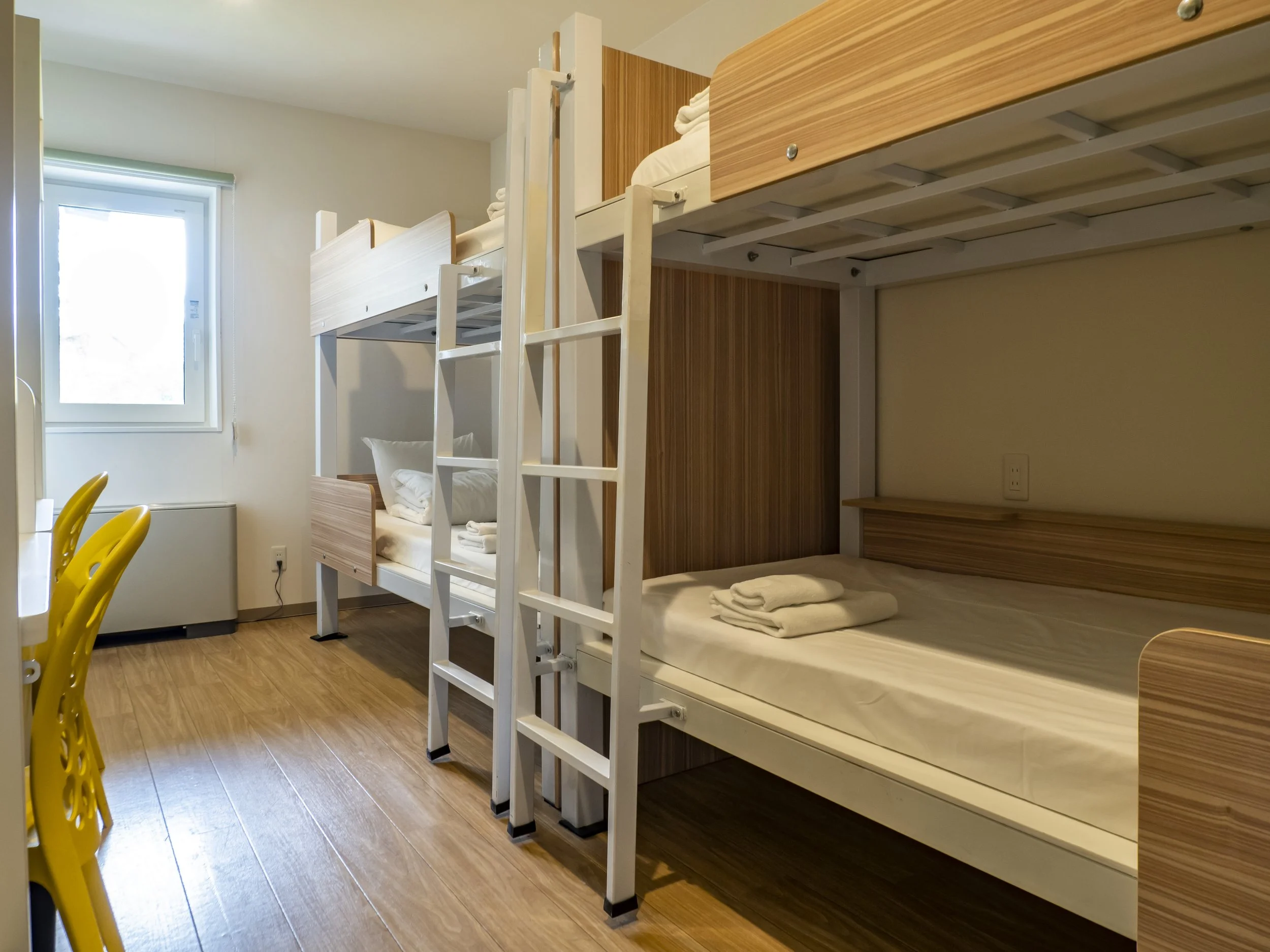 My Ecolodge, Hirafu, Niseko — A dormitory room with a bunk bed, a window, and a yellow chair.